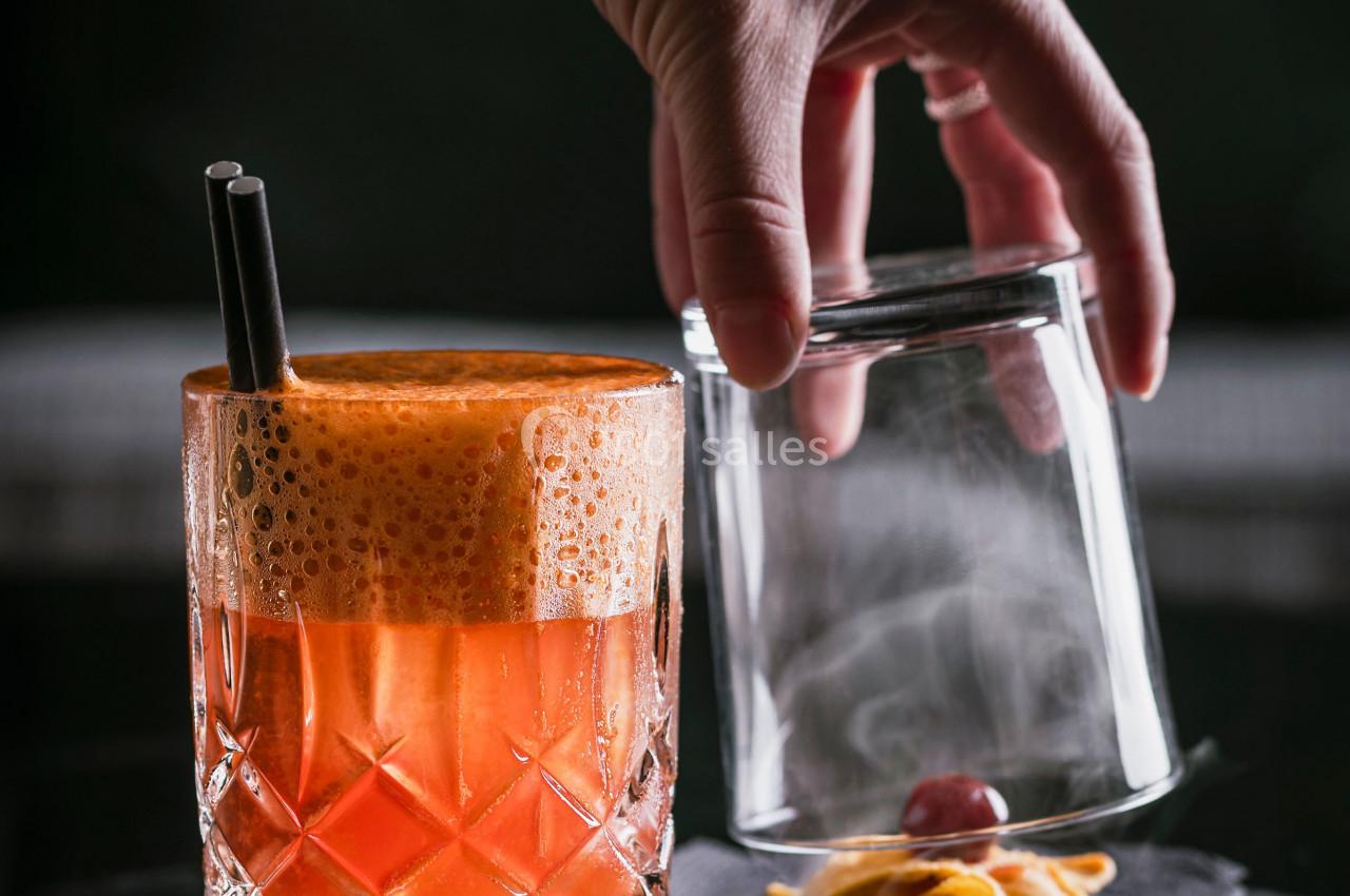 Main déposant un verre fumé à côté d'un cocktail orange mousseux, décoré d'une cerise et d'un zeste d'agrume.