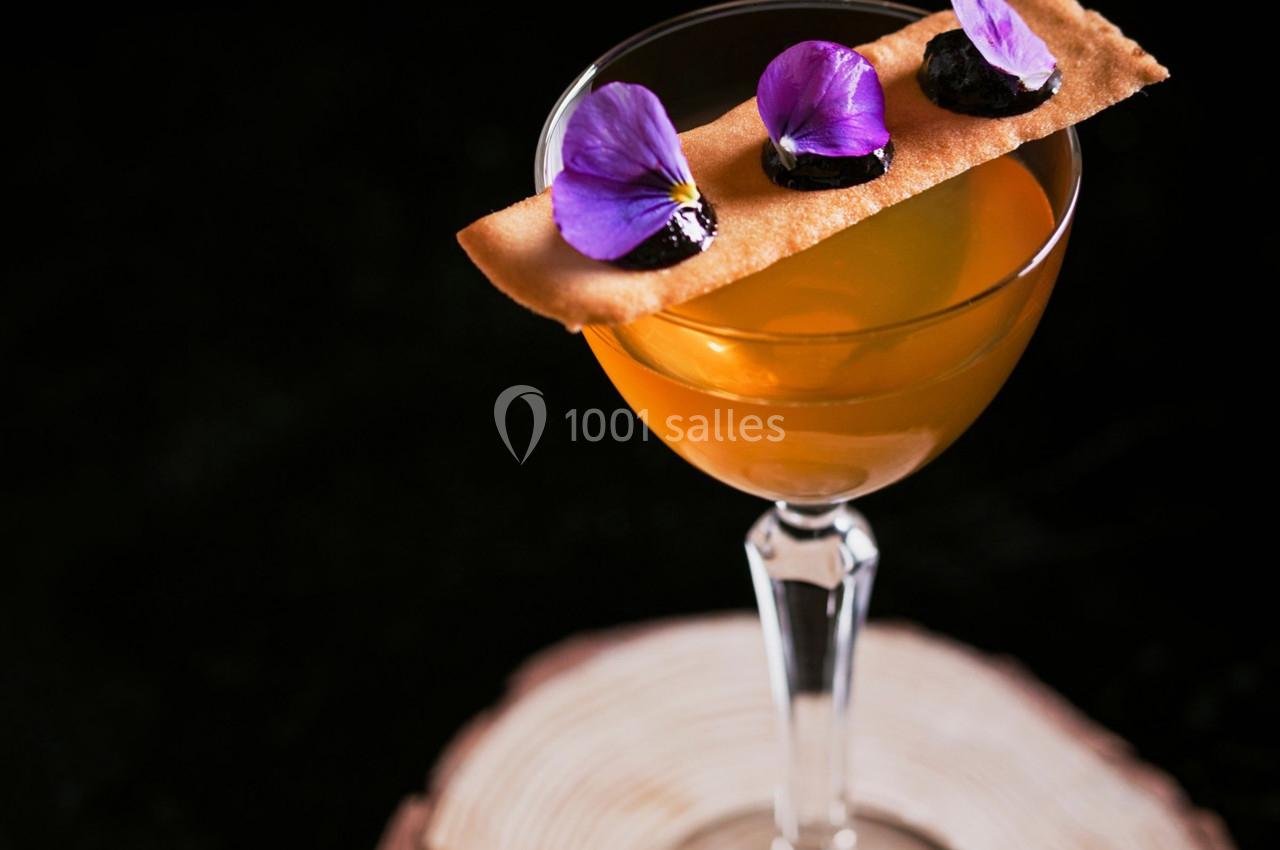 Verre à cocktail orange décoré d'une fine tuile, de fleurs violettes et de touches sombres, posé sur un support en bois.