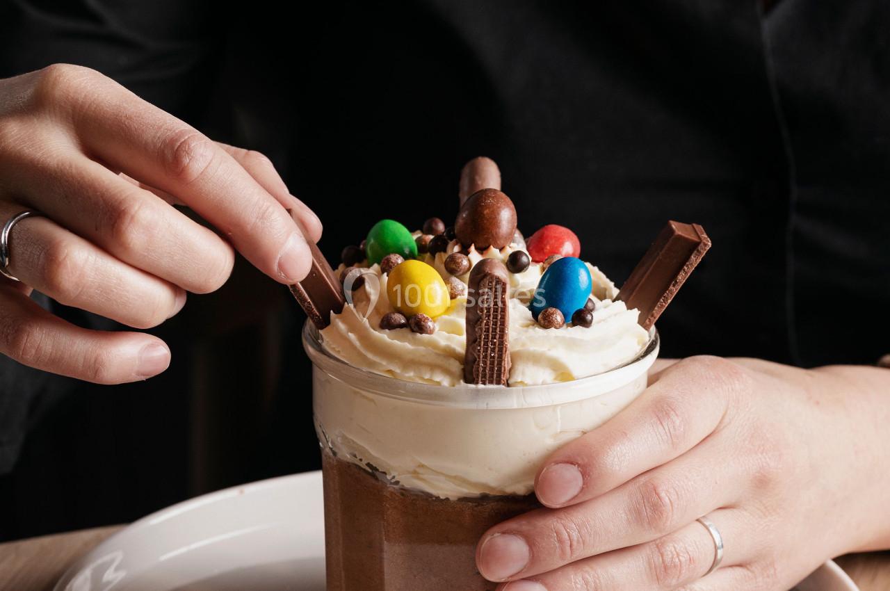 Une personne tient un verre de dessert chocolaté garni de crème fouettée, bonbons colorés et morceaux de chocolat.