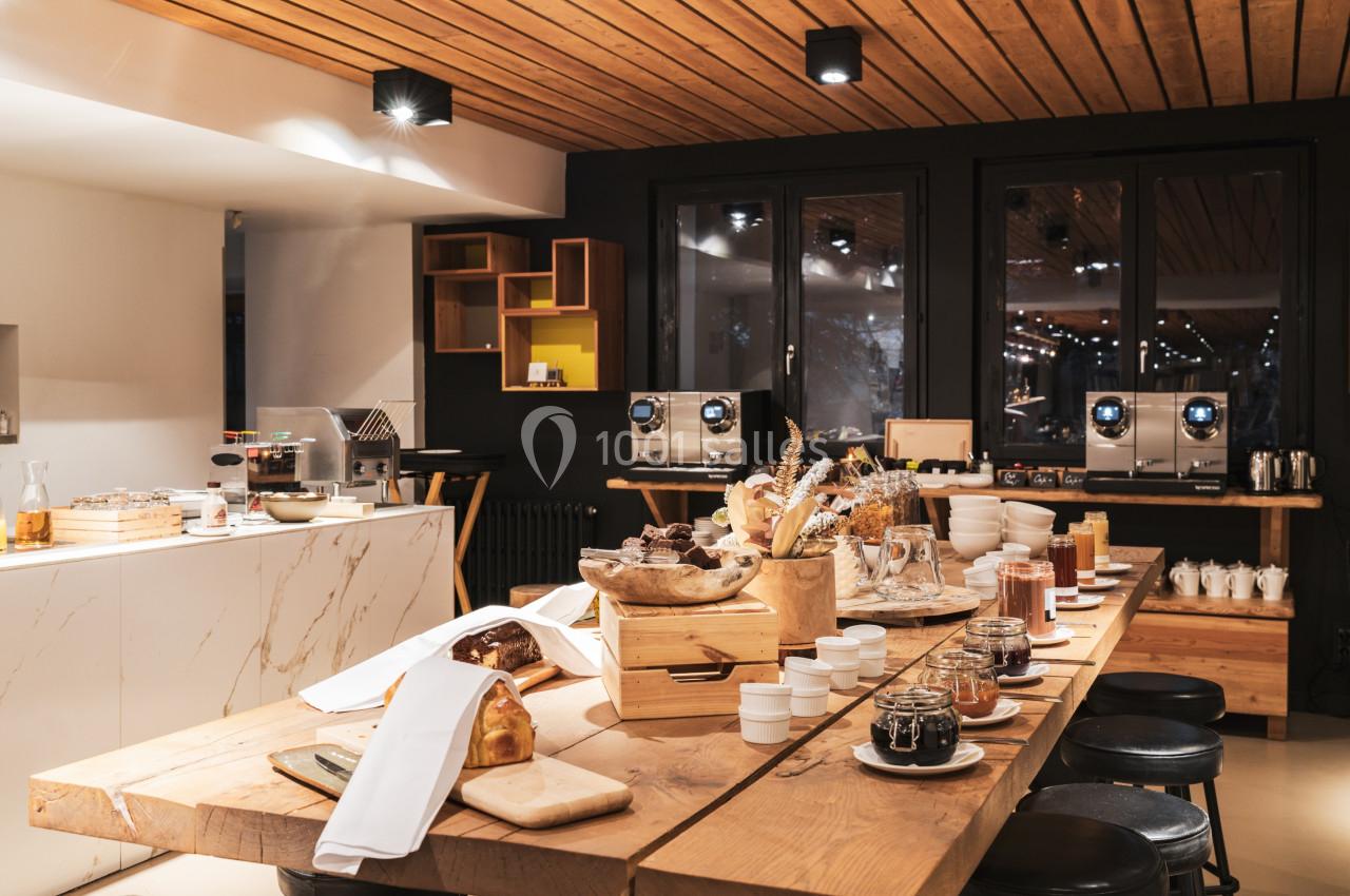 Table en bois garnie de pains, confitures et boissons dans une salle lumineuse avec machines à café en arrière-plan.