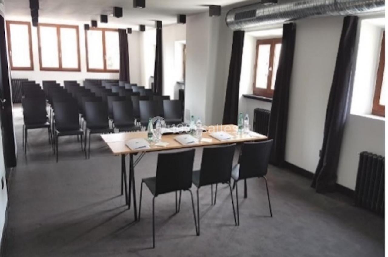Salle de réunion lumineuse avec rangées de chaises noires, tables équipées de bouteilles d'eau et documents.