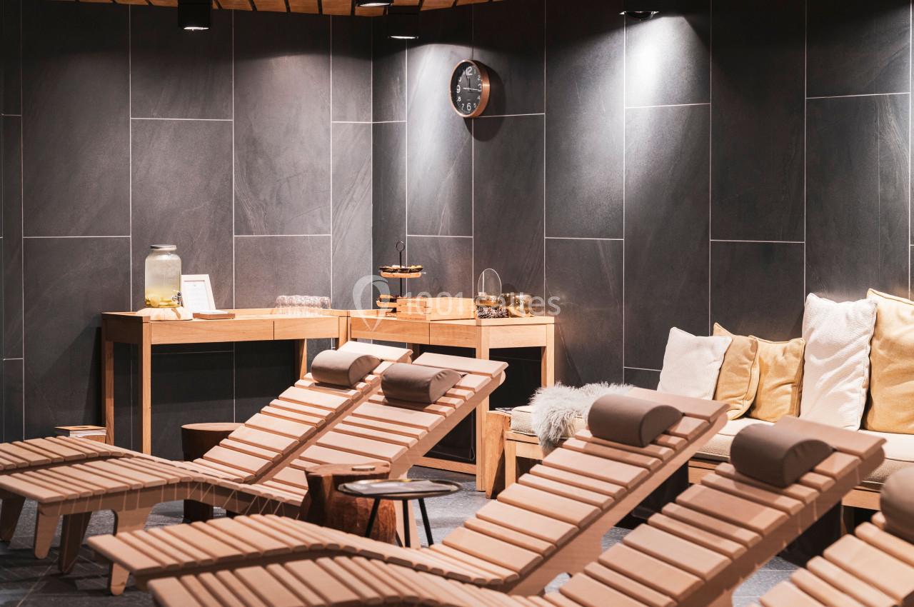 Salle de détente avec chaises longues en bois, coussins, éclairage tamisé et table avec boissons et collations.