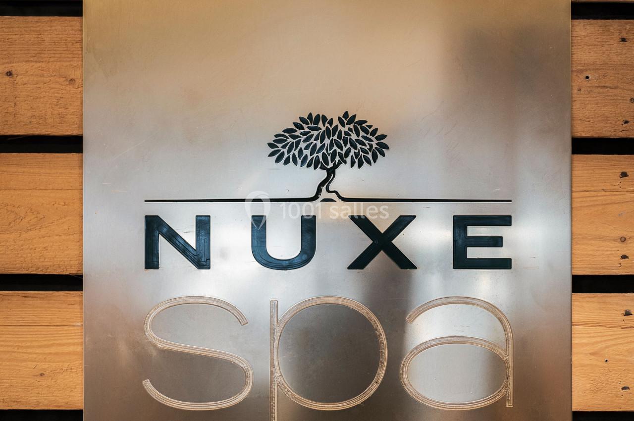 Plaque métallique avec le logo ’Nuxe Spa Paris’ gravé, fixée sur un mur en bois.