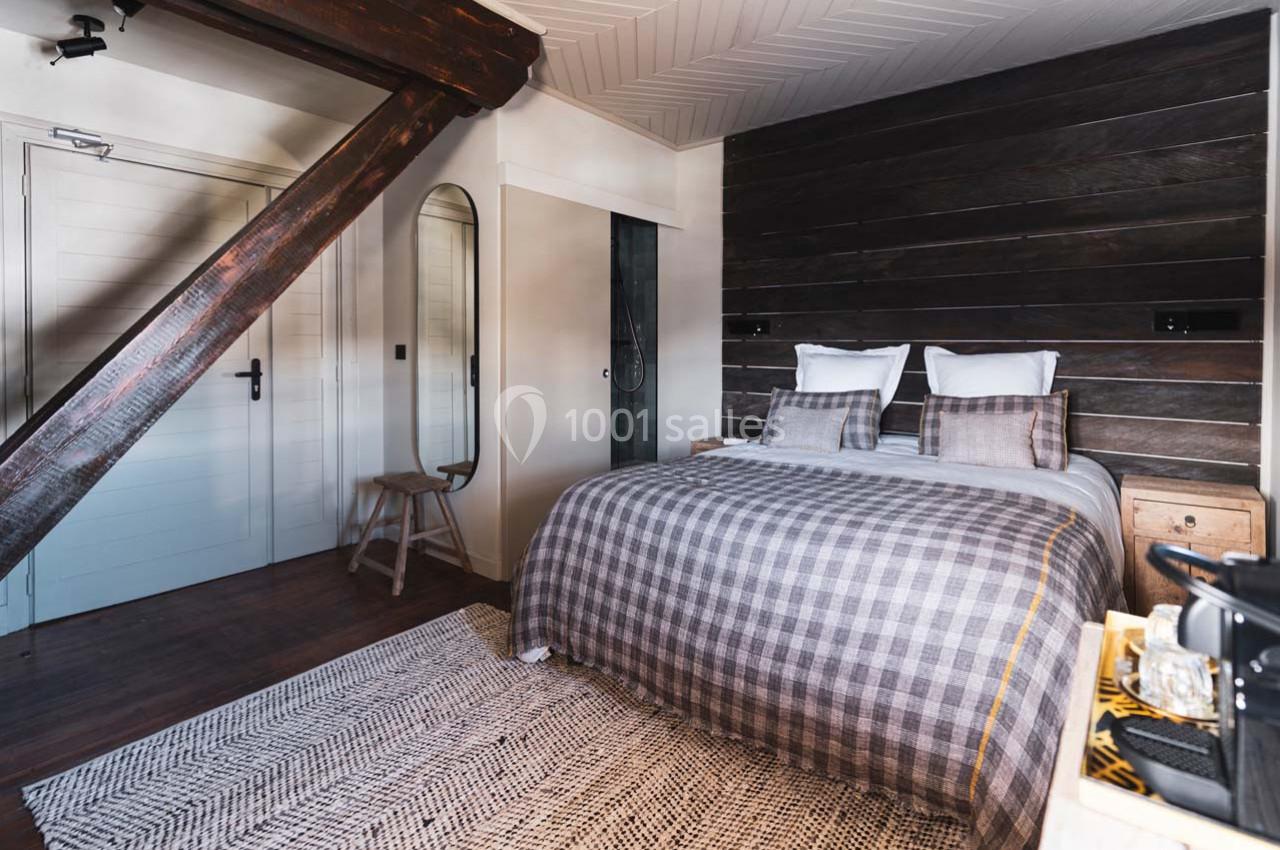 Chambre avec lit double, plaid à carreaux, mur en bois sombre, miroir sur pied et poutre apparente.