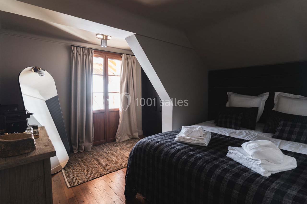 Chambre lumineuse avec lit double, linge plié, grand miroir, fenêtre avec rideaux et sol en bois.