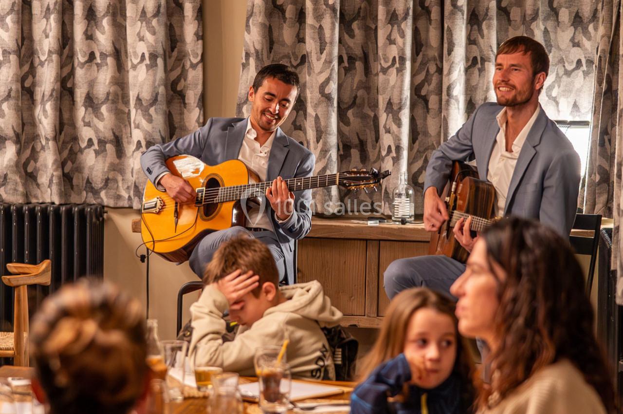 Deux musiciens en costume jouent de la guitare devant un petit public dans un cadre intérieur chaleureux.