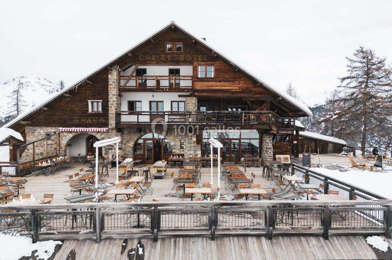 Chalet en bois avec terrasse aménagée, entouré de neige et de montagnes, sous un ciel nuageux.