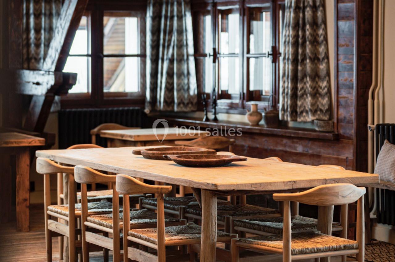 Salle à manger rustique avec une grande table en bois entourée de chaises, éclairée par des fenêtres avec rideaux à motifs.