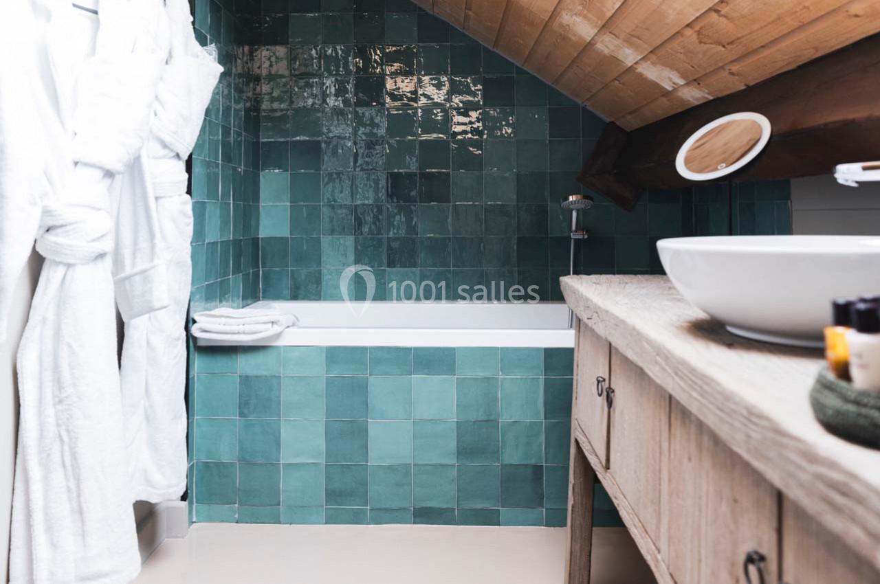 Salle de bain sous combles avec mur en carreaux verts, baignoire, lavabo sur meuble en bois et peignoirs accrochés.