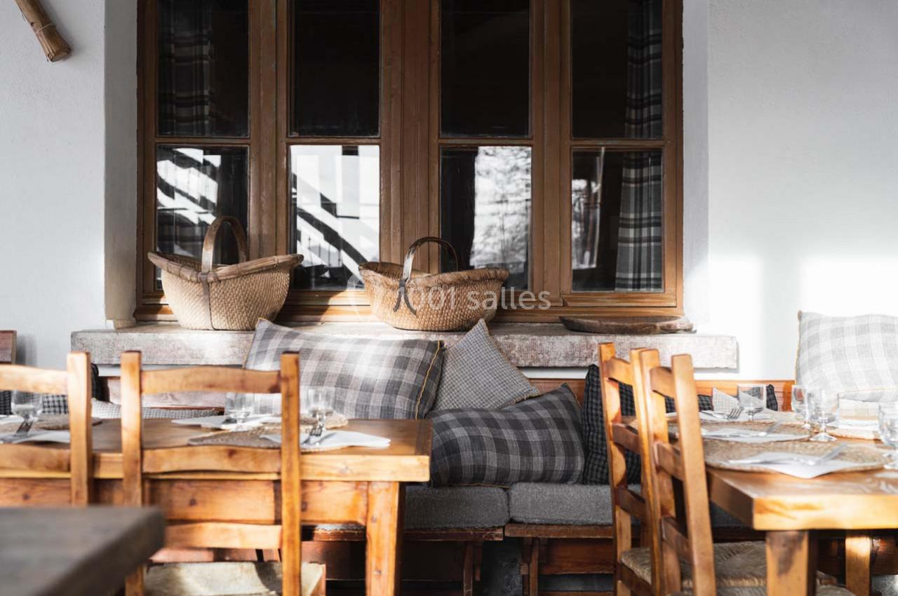 Tables en bois dressées près d'une banquette avec coussins et paniers en osier, devant une fenêtre à carreaux.