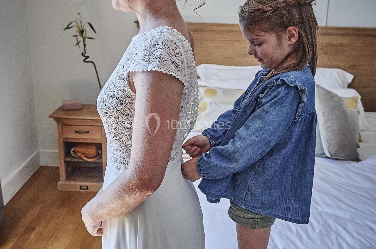 Une petite fille aide une femme à fermer sa robe blanche dans une chambre lumineuse.