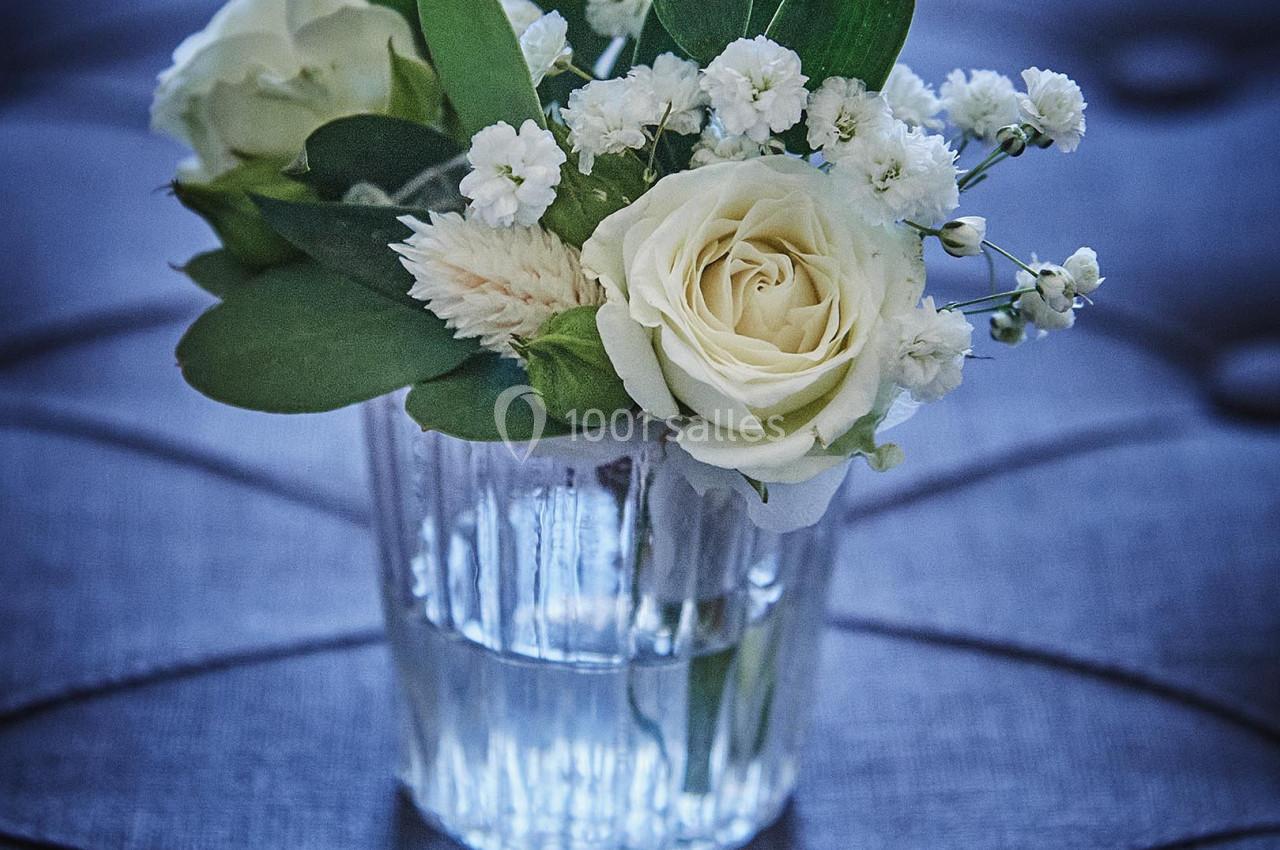 Petit bouquet de fleurs blanches et feuillage vert dans un vase en verre, posé sur un tissu matelassé gris.
