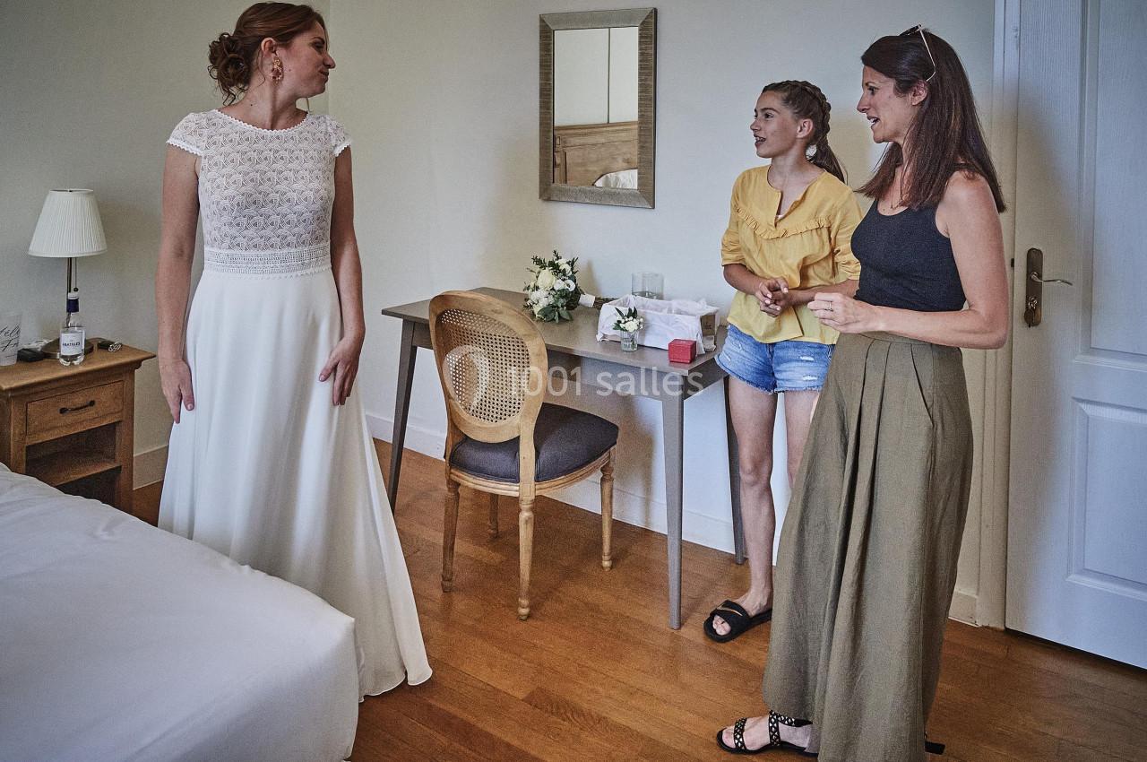 Une femme en robe de mariée discute avec deux femmes dans une chambre lumineuse avec un bureau en arrière-plan.