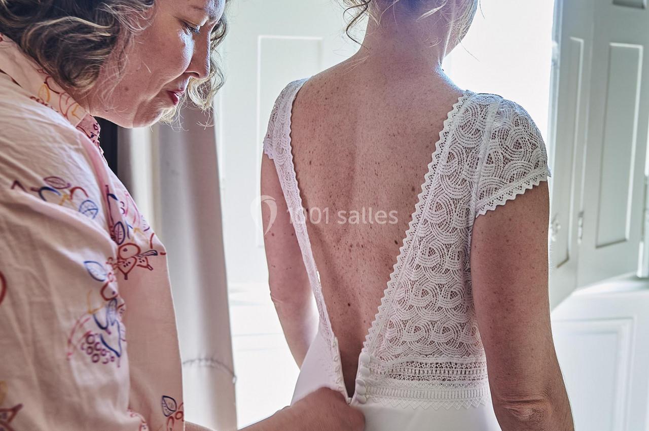 Une femme aide une mariée à fermer sa robe en dentelle blanche dans une pièce lumineuse avec des volets ouverts.