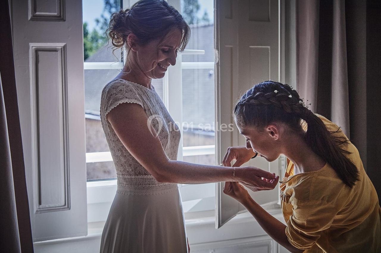 Une femme en robe blanche sourit tandis qu'une autre attache un bracelet à son poignet près d'une fenêtre ouverte.
