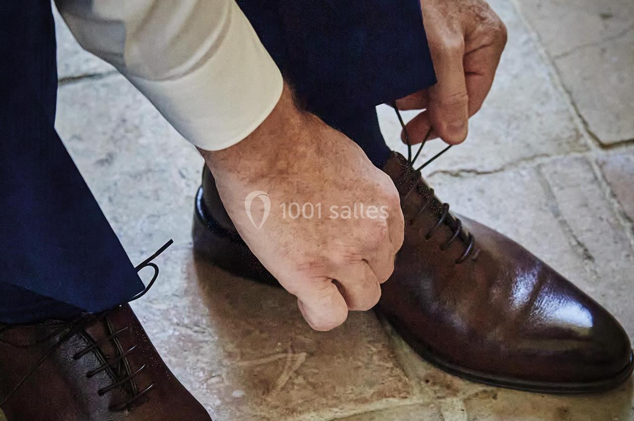 Un homme en costume noue les lacets de ses chaussures en cuir marron sur un sol en carrelage.