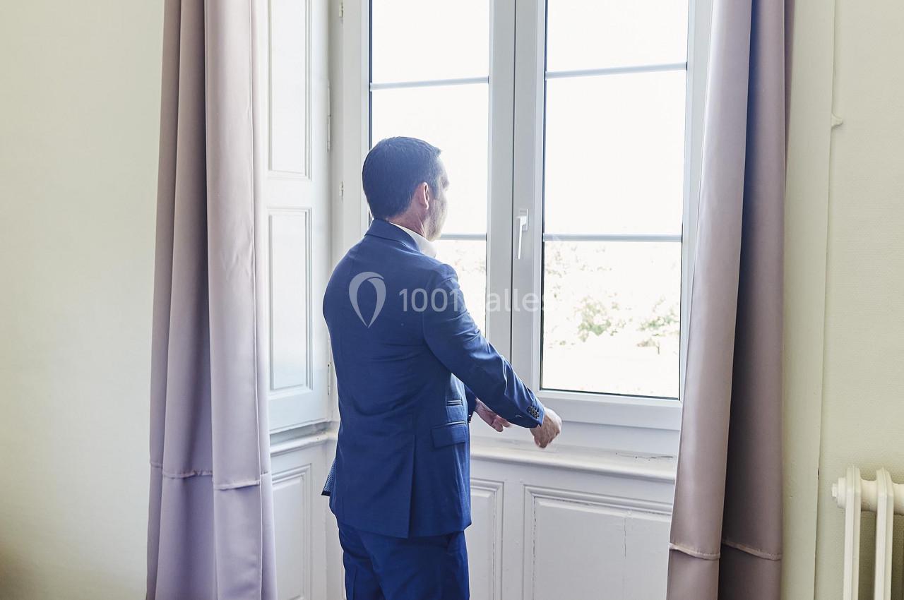 Un homme en costume regarde par une grande fenêtre dans une pièce lumineuse avec des rideaux et un sol en carreaux.