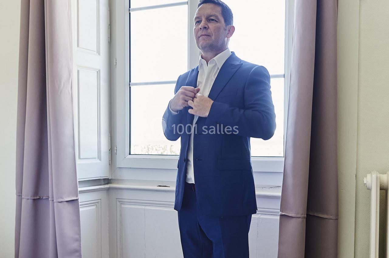 Homme en costume bleu ajustant sa chemise devant une grande fenêtre dans une pièce lumineuse.