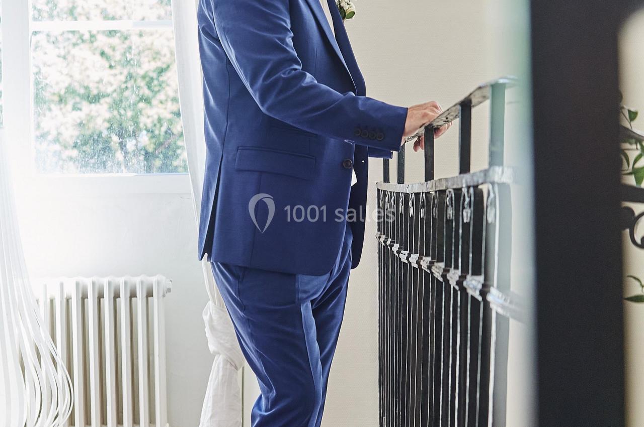 Homme en costume bleu debout près d'une rambarde, dans une pièce lumineuse avec un sol en terre cuite.