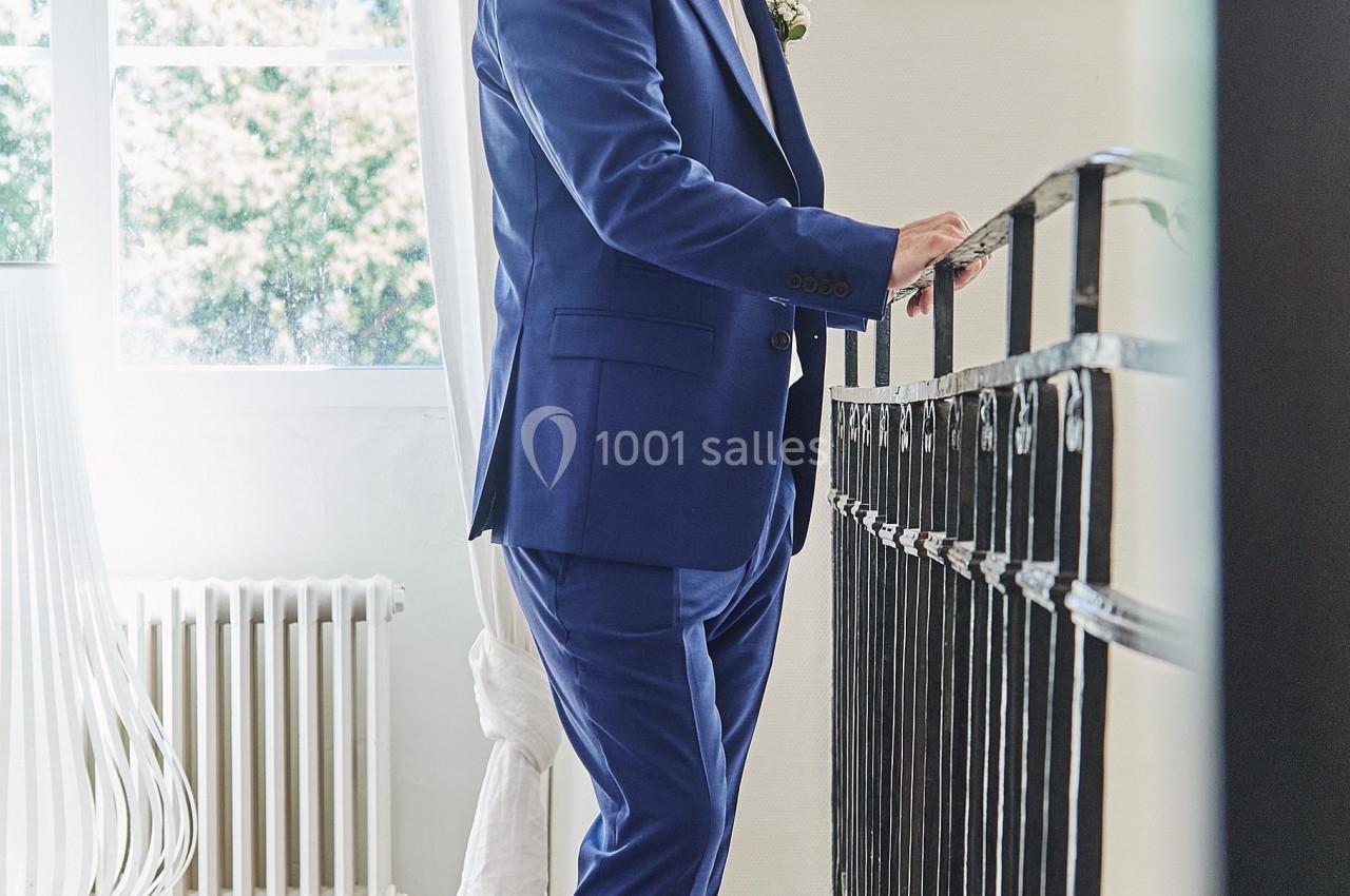 Homme en costume bleu appuyé sur une rambarde dans une pièce lumineuse avec une fenêtre et un décor épuré.