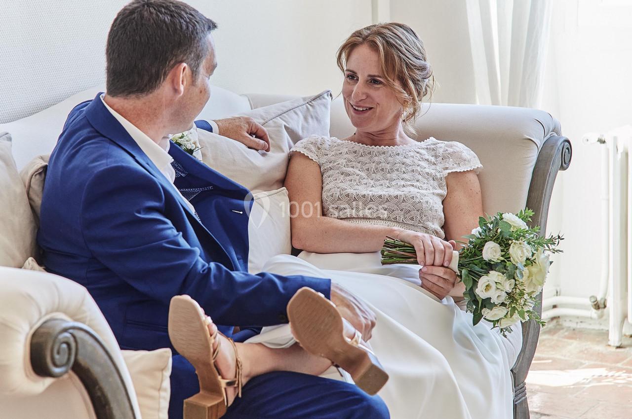 Un couple en tenue de mariage discute sur un canapé, la mariée tenant un bouquet de fleurs blanches.