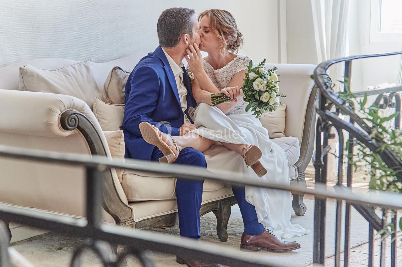 Un couple de mariés s'embrasse tendrement sur un canapé, la mariée tenant un bouquet de fleurs blanches.
