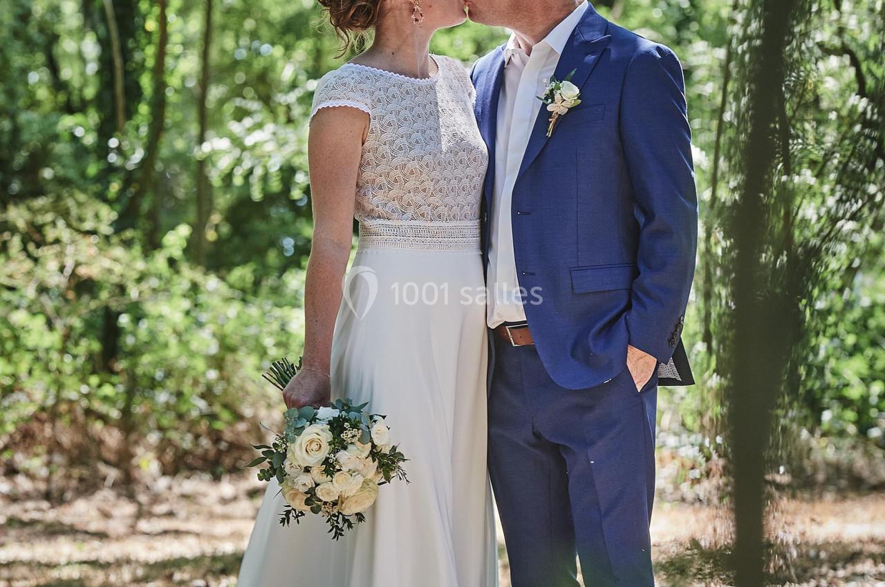 Un couple en tenue de mariage s'embrasse dans une clairière boisée, sous une lumière naturelle.