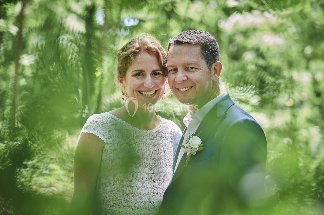 Un couple souriant en tenue de mariage, entouré de verdure et baigné par une lumière naturelle.