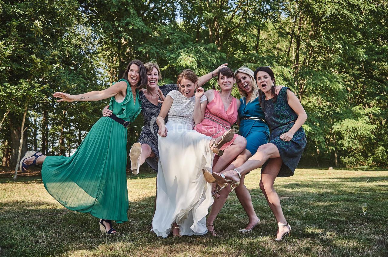 Six femmes souriantes en tenue de fête lèvent une jambe en riant, debout sur une pelouse devant des arbres.