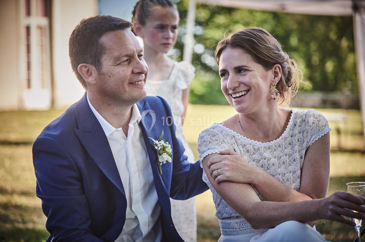 Un couple souriant assis à l'extérieur lors d'une réception, avec une jeune fille en arrière-plan.
