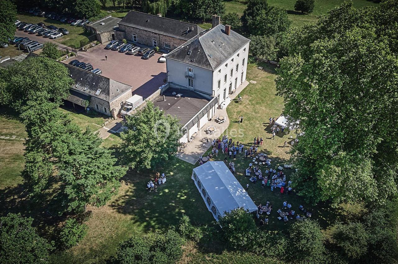 Vue aérienne d'un domaine avec un bâtiment principal, des arbres, un parking et une réception sous une tente blanche.