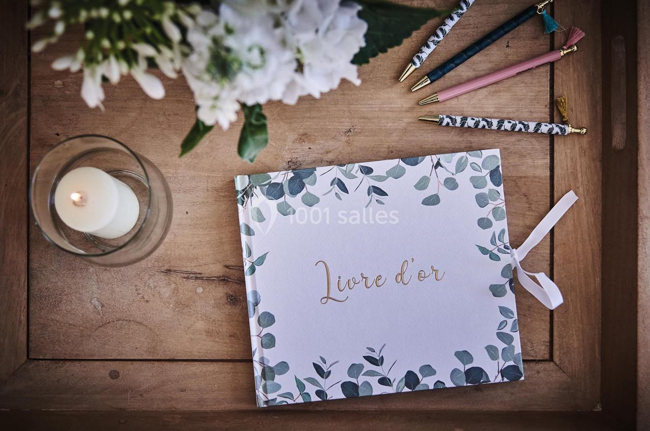 Livre d'or décoré de motifs végétaux posé sur une table en bois avec une bougie, des fleurs et des stylos colorés.