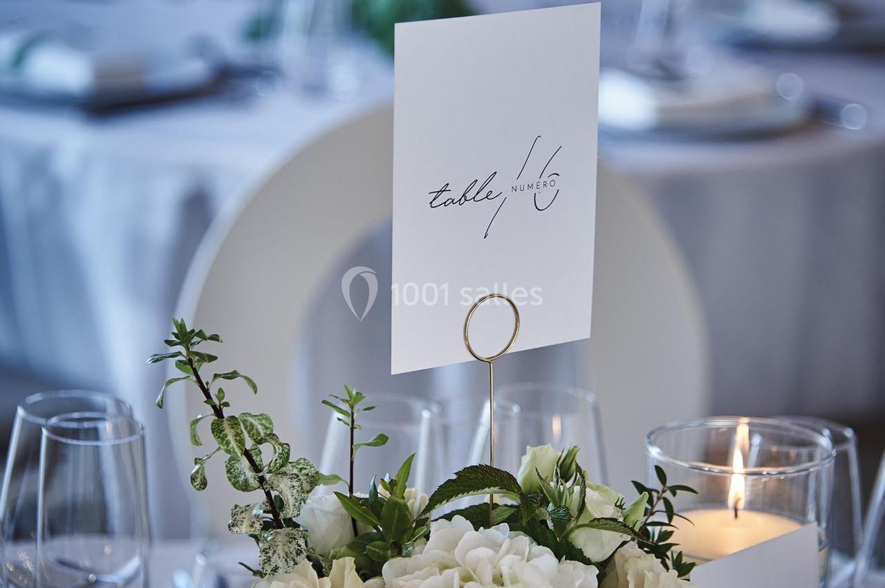 Centre de table avec fleurs blanches, bougies allumées et carte de table sur une pince dorée, dans une salle de réception.