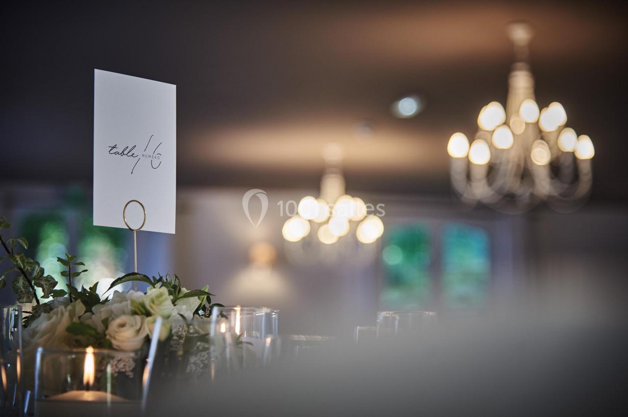 Carte de table numérotée entourée de bougies et fleurs, avec des lustres flous en arrière-plan.