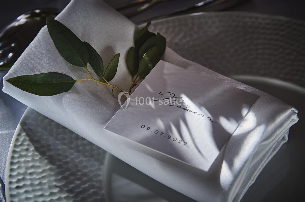 Serviette blanche pliée avec une branche d'eucalyptus, posée sur une assiette texturée, accompagnée d'une carte marquée ’09…