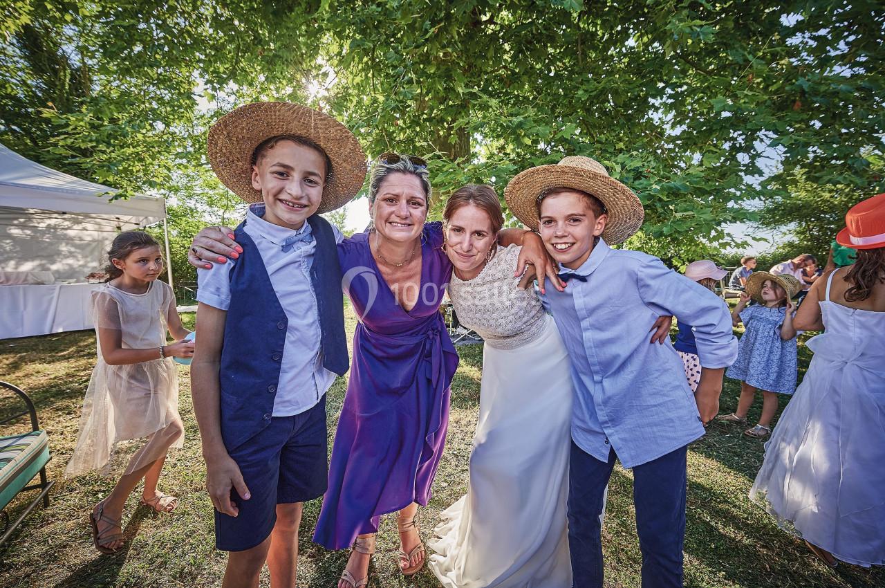 Deux enfants et deux femmes, dont une en robe de mariée, posent souriants sous des arbres lors d'un événement en extérieur.
