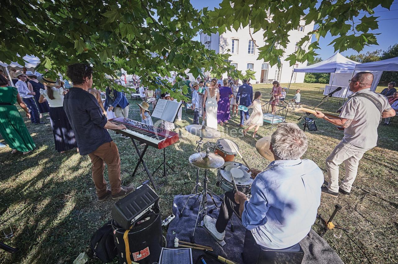 Musiciens jouant en plein air sous un arbre lors d'un événement avec des spectateurs en arrière-plan.