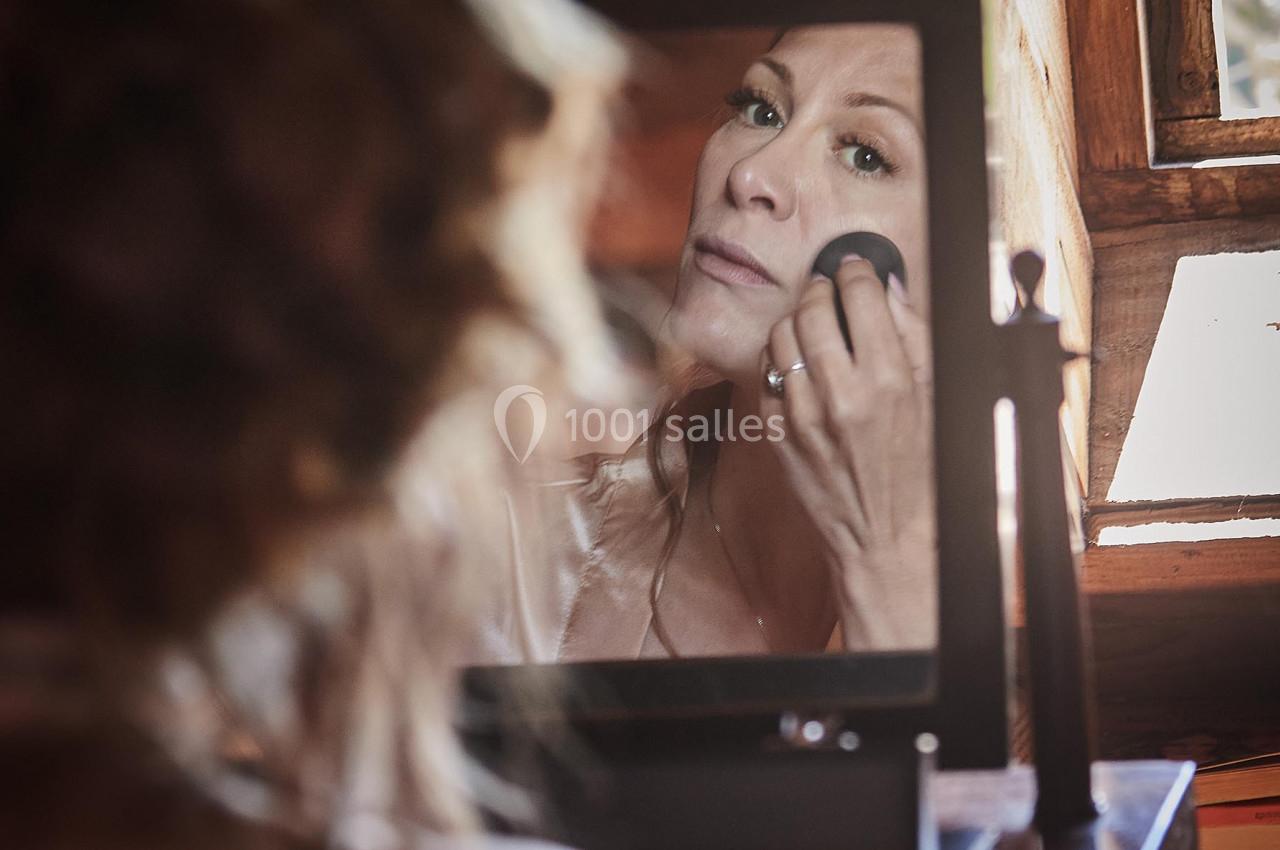 Femme appliquant du maquillage devant un miroir dans une pièce en bois éclairée par la lumière naturelle.