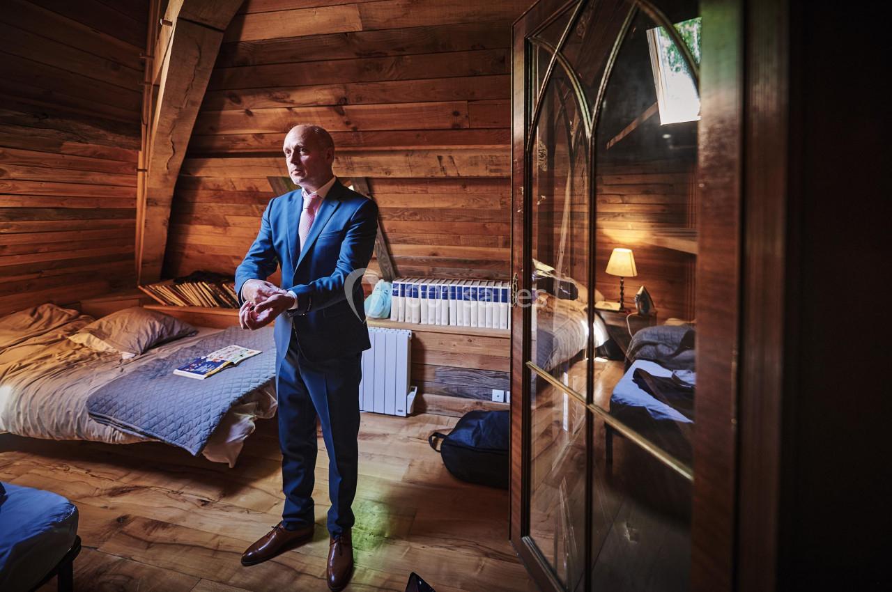 Un homme en costume ajuste ses manches dans une chambre en bois avec un lit, une lampe et des livres.