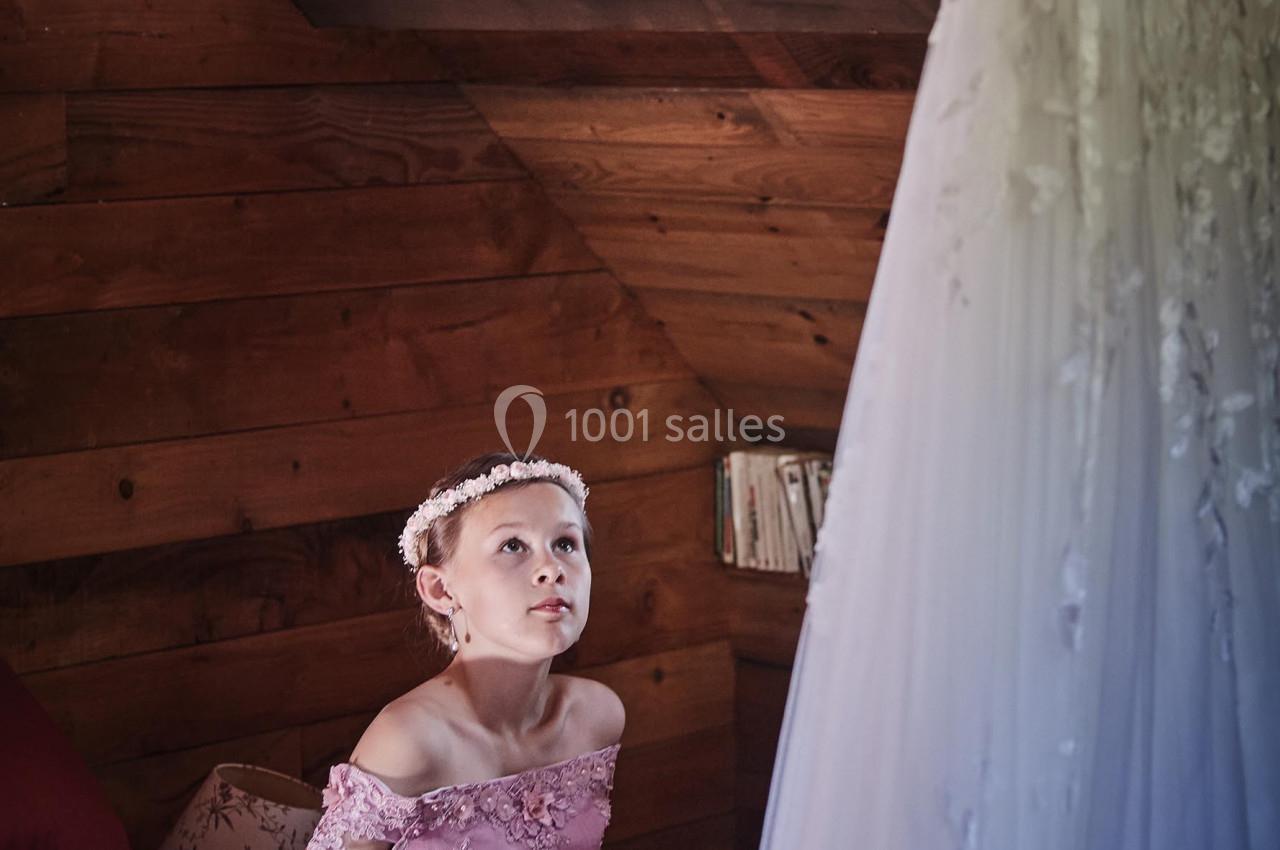 Une jeune fille en robe rose regarde une robe de mariée suspendue dans une pièce en bois.