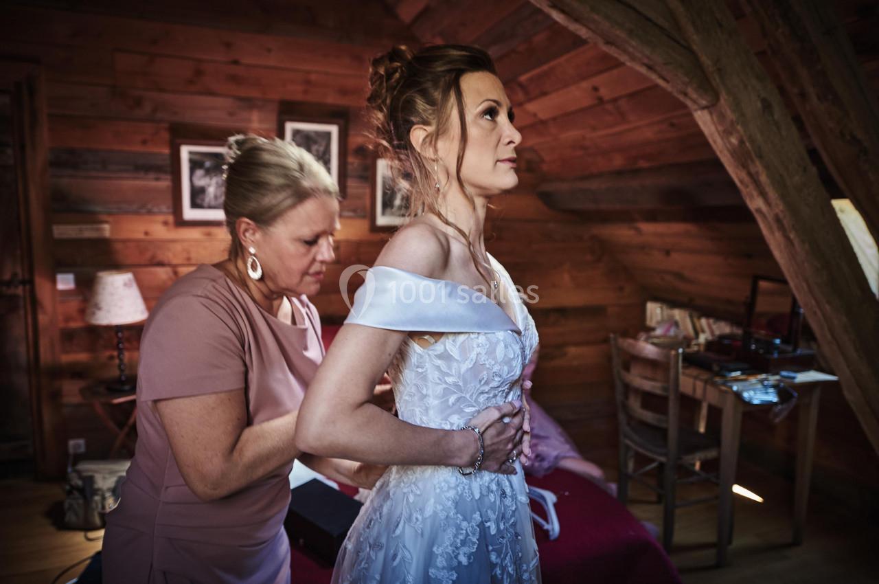 Une femme aide une mariée à ajuster sa robe dans une pièce en bois au décor rustique.