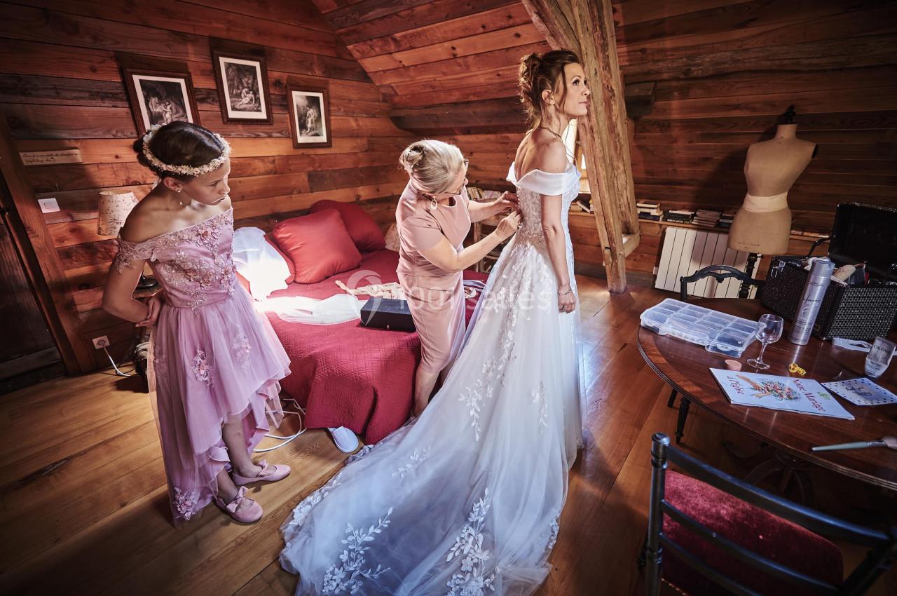 Une mariée en robe blanche est ajustée par une personne, accompagnée d'une demoiselle d'honneur dans une chambre en bois.