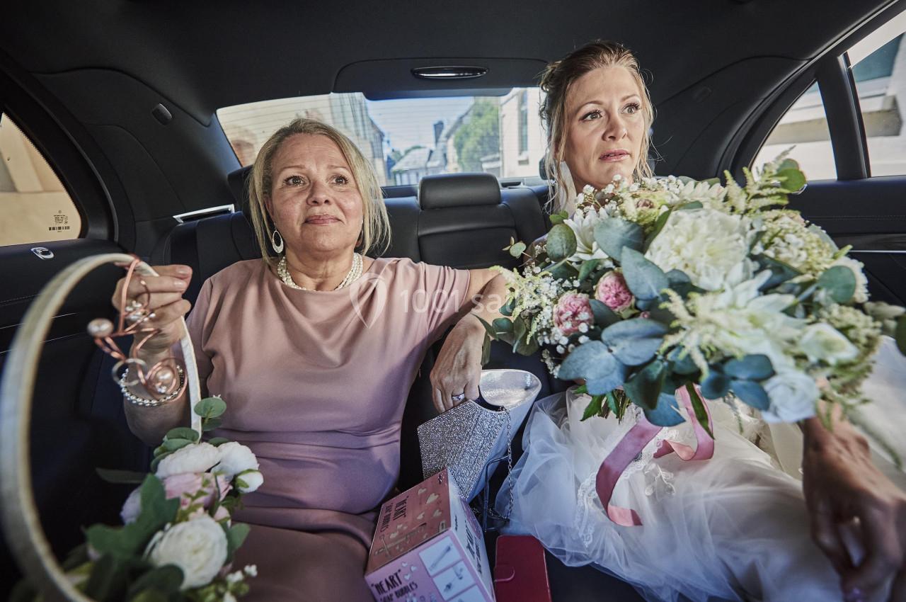 Deux femmes assises dans une voiture, l'une en robe de mariée tenant un bouquet, l'autre en tenue élégante avec des…