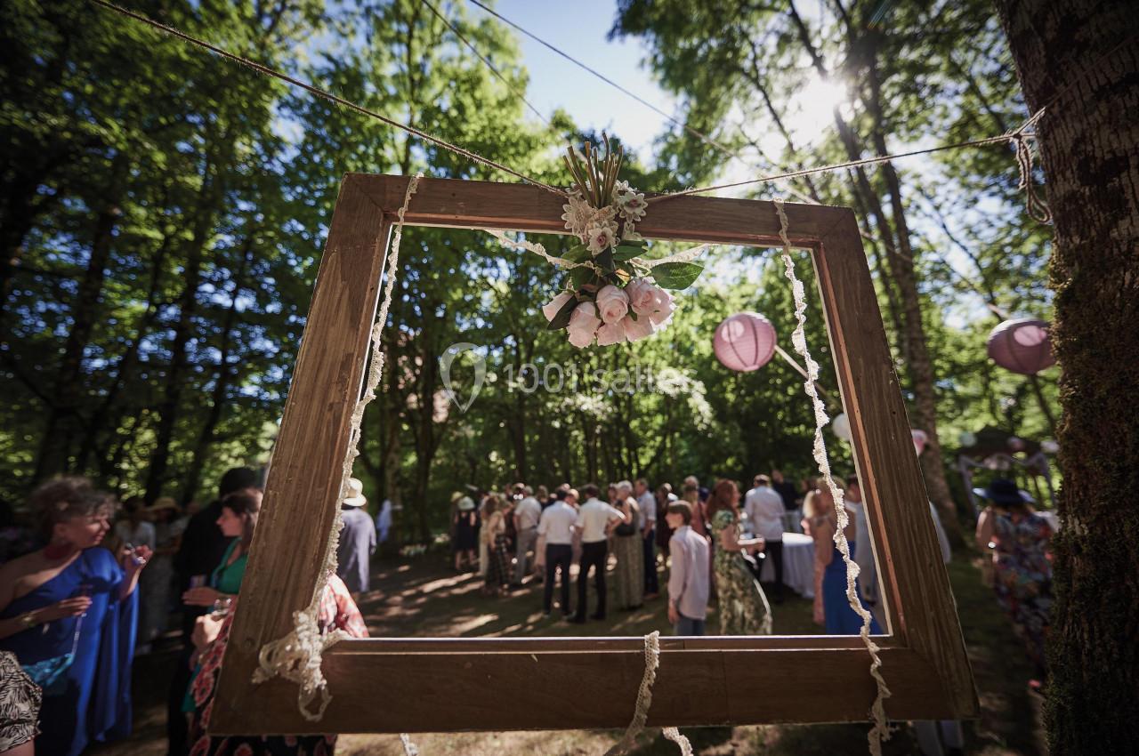 Cadre en bois décoré de fleurs suspendu dans une forêt lors d'un événement en plein air avec des invités en arrière-plan.