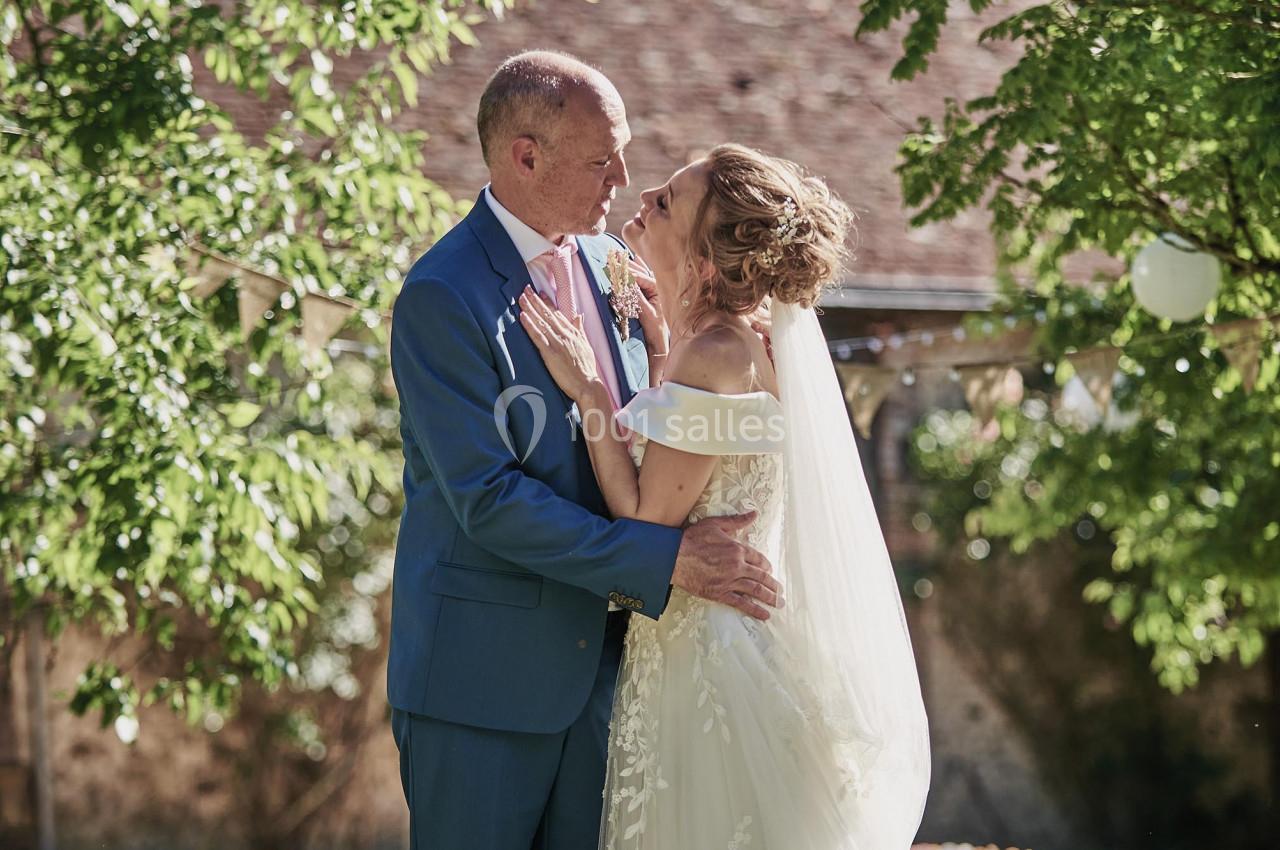 Un couple de mariés se tient debout dans un jardin ensoleillé, entouré d'arbres et de décorations colorées.