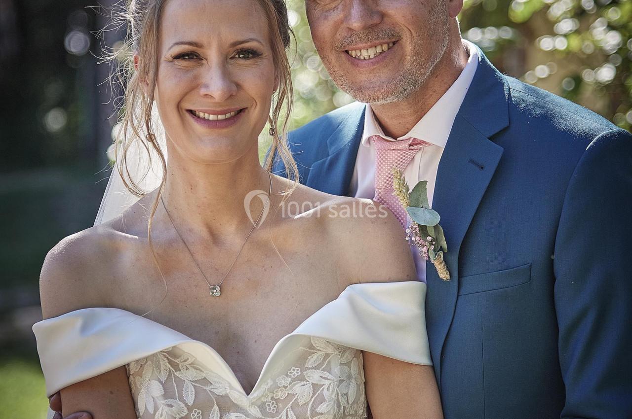 Un couple souriant en tenue de mariage pose dans un jardin verdoyant sous une lumière naturelle.