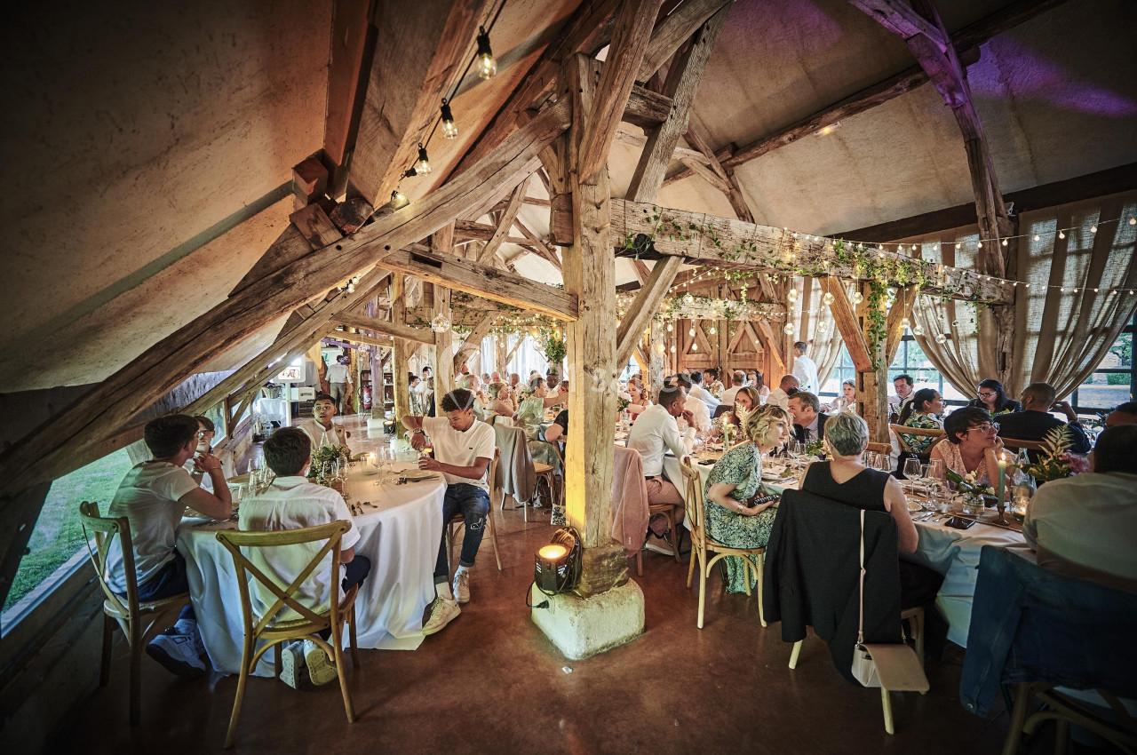 Salle de réception rustique avec poutres apparentes, décorée de guirlandes lumineuses, accueillant des convives à table.
