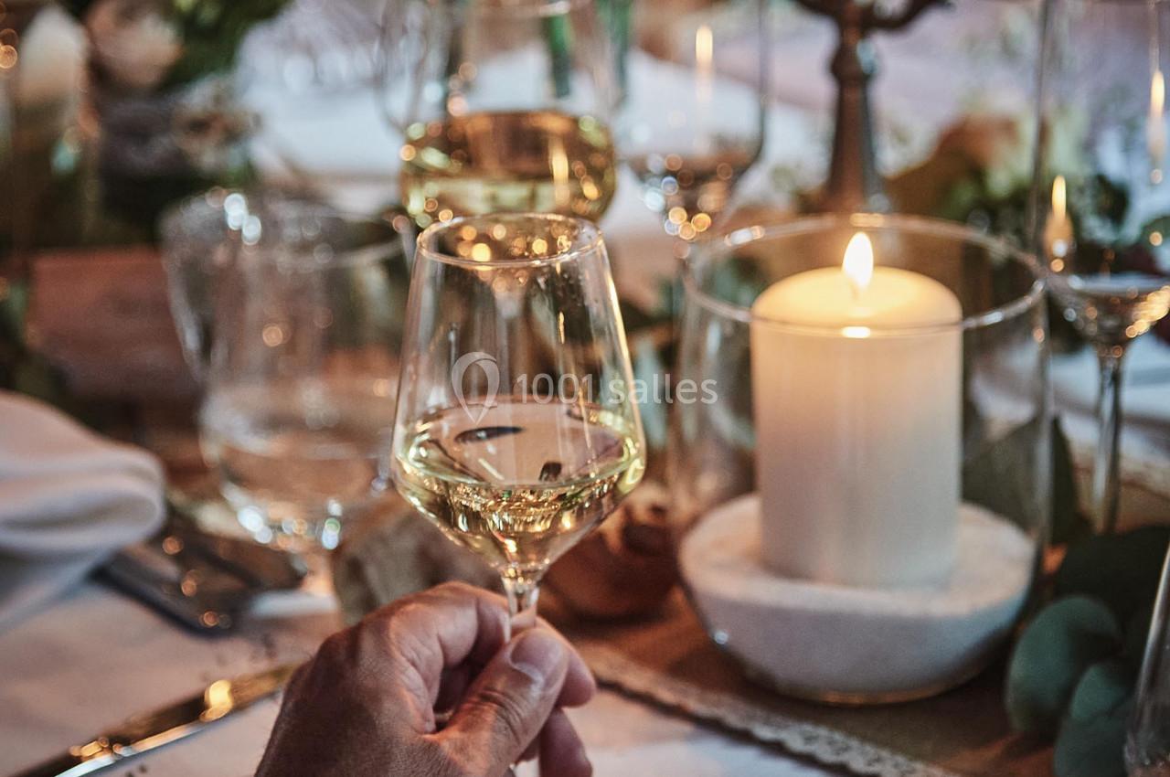 Main tenant un verre de vin blanc sur une table élégamment dressée avec bougies et décorations florales.