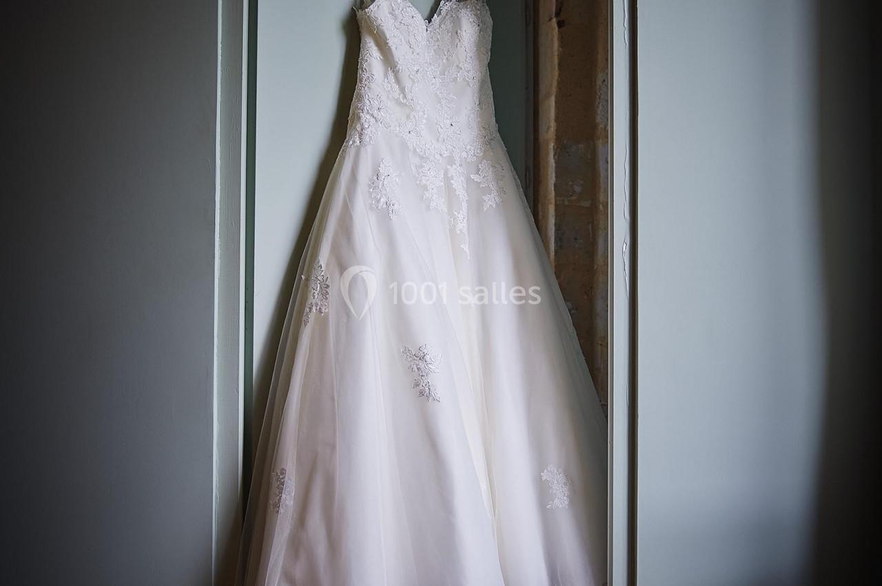 Robe de mariée blanche en dentelle suspendue à une porte dans une pièce éclairée par une lumière douce.