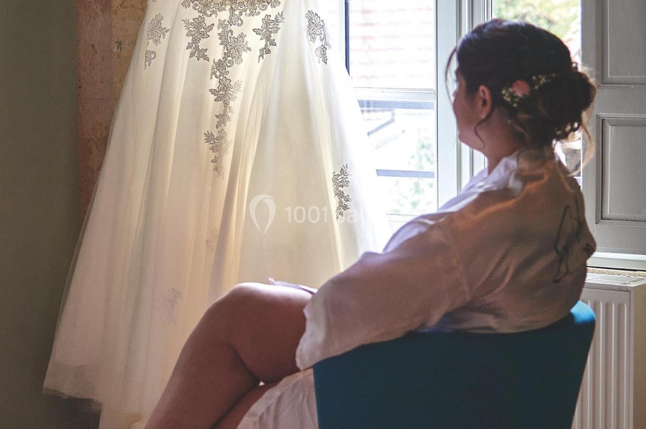 Une femme assise observe une robe de mariée suspendue devant une fenêtre lumineuse.